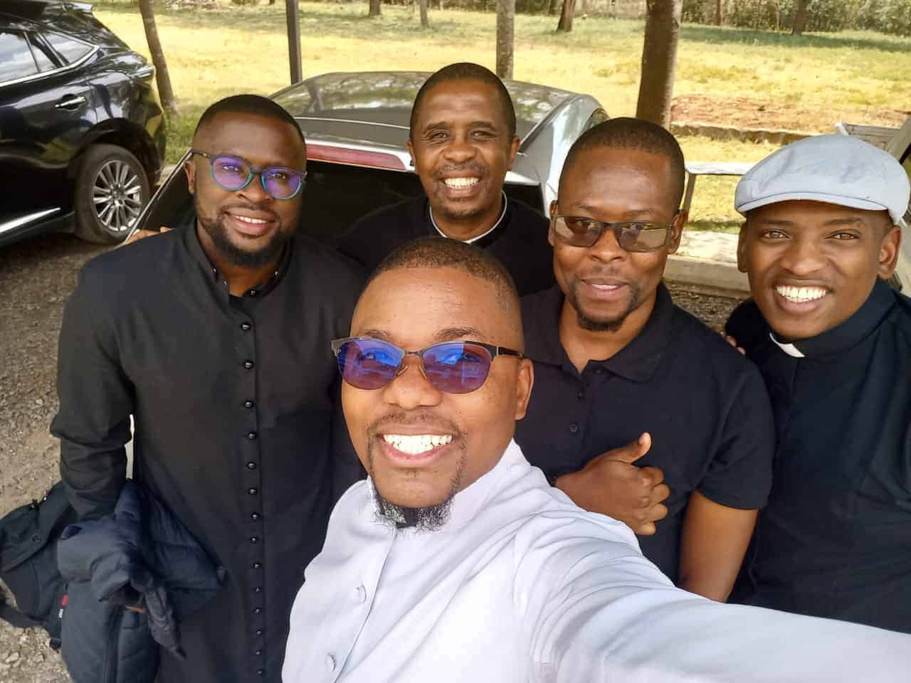 Five smiling men stand outdoors, posing for a selfie. They are dressed in dark and light clothing, with some wearing clerical collars. Trees, parked cars, and grass can be seen in the background.